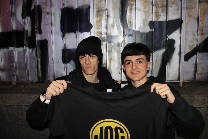 Dos personas sostienen una camiseta negra con el logo amarillo de JOC District ante una pared con grafitis.